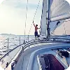 海の上を航行するヨットの甲板に立つ人物が、帆を支えながら景色を楽しんでいる様子